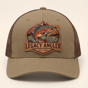 May include: A tan trucker cap with a brown mesh back and a leather-like patch on the front. The patch features a colourful illustration of a jumping trout, mountains, and the text LEGACY ANGLER EST. 1983 in a dark brown, bold, serif font.
