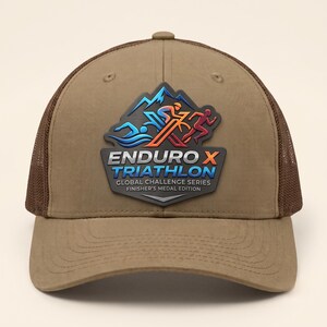 May include: A tan baseball cap with brown mesh back panels and a front patch featuring a mountain graphic, figures swimming, cycling, and running, and the text ENDURO X TRIATHLON GLOBAL CHALLENGE SERIES FINISHER'S MEDAL EDITION in blue and white.