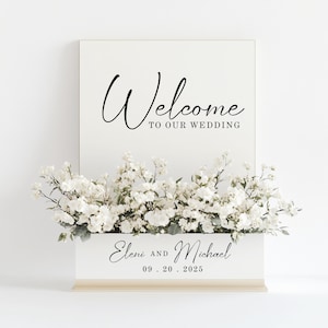 May include: A white wedding welcome sign with the word "Welcome" in elegant black script. Below, it reads "TO OUR WEDDING". The sign is decorated with white flowers and the names "Eleni and Michael" with the date 09.20.2025.