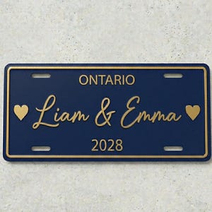 Peut inclure: Plaque d'immatriculation bleu marine avec bordure et texte dorés. On peut lire "ONTARIO" en haut, avec "Liam & Emma" en cursive et "2028" en dessous. Des cœurs dorés encadrent les noms.