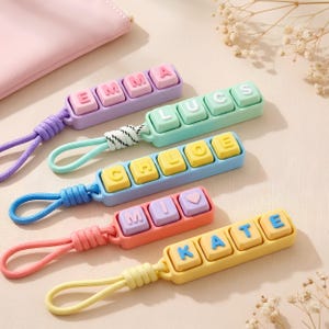 May include: Five rectangular keychains with colourful plastic bases and raised square letter keys. The keychains feature the names EMMA, LUCS, CHLOE, MIO with a heart symbol, and KATE, each attached to a matching braided cord loop for easy attachment.