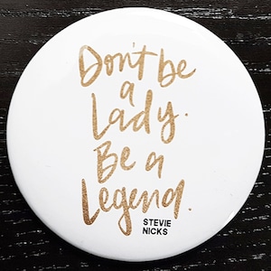 Pu&ograve; includere: Bottone rotondo bianco con scritta dorata "Don't be a lady. Be a legend." con il testo "Stevie Nicks" in lettere pi&ugrave; piccole sotto.