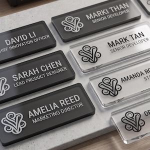 May include: A collection of rectangular name badges in black and clear acrylic. Each badge features a stylised floral logo on the left and printed text including names like Marki Than, David Li, Sarah Chen, and Amelia Reed with job titles.