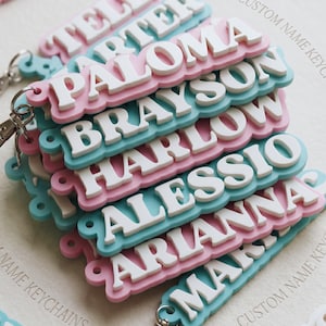 May include: A stack of custom name keychains featuring white raised lettering on alternating pastel pink and light blue scalloped bases. Each keychain includes a metal lobster clasp attachment and displays names such as Paloma, Brayson, Harlow, Alessio, and Arianna.