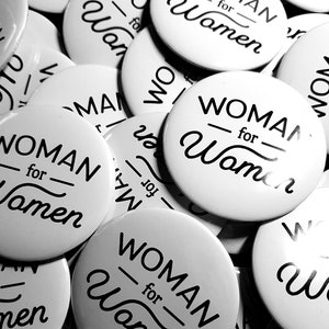Pu&ograve; includere: Primo piano di diversi bottoni rotondi bianchi con testo nero che dice "WOMAN for Women".