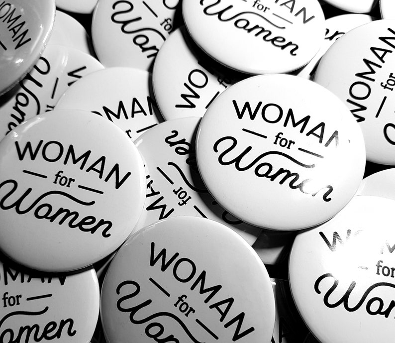 Pu&ograve; includere: Primo piano di diversi bottoni rotondi bianchi con testo nero che dice "WOMAN for Women".