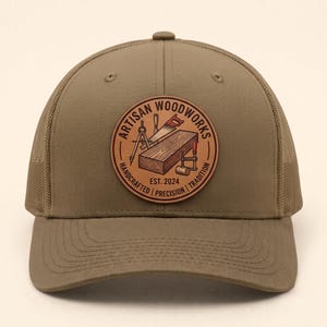 May include: A khaki-coloured baseball cap with a mesh back and a circular leather patch on the front. The patch features a wood grain illustration, a saw, a compass, and a clamp, with the text ARTISAN WOODWORKS EST. 2024 HANDCRAFTED PRECISION TRADITION.