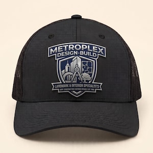 May include: A black baseball cap with a mesh back and a front patch featuring the text METROPLEX DESIGN-BUILD LANDMARK & INTERIOR SPECIALISTS CORP. CONSTRUCTION | OUTDOOR SPACES | EST. 1998, alongside a shield graphic with a compass and building.