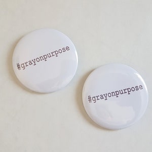 Pu&ograve; includere: Due bottoni rotondi bianchi con il testo "#crayonpurpose" stampato in grigio.