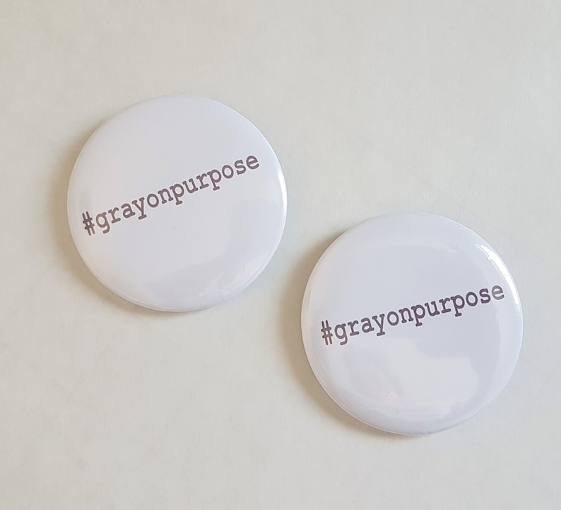 Pu&ograve; includere: Due bottoni rotondi bianchi con il testo "#crayonpurpose" stampato in grigio.