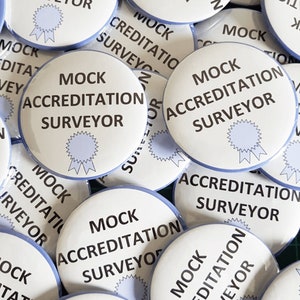 Pu&ograve; includere: Primo piano di diversi bottoni rotondi bianchi con bordo blu. I bottoni hanno il testo "MOCK ACCREDITATION SURVEYOR" stampato in nero. Ogni bottone presenta un nastro blu con una stella.