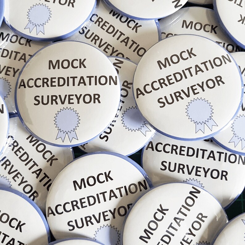 Pu&ograve; includere: Primo piano di diversi bottoni rotondi bianchi con bordo blu. I bottoni hanno il testo "MOCK ACCREDITATION SURVEYOR" stampato in nero. Ogni bottone presenta un nastro blu con una stella.