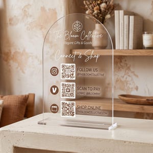 Peut inclure: Un panneau en acrylique transparent en forme d'arche avec du texte et des graphiques imprimés en blanc. Le panneau affiche le logo The Bloom Collective, Connect & Shop, et trois codes QR avec des icônes pour Instagram, Venmo et un site web.
