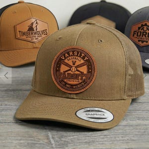 May include: Several baseball caps with leather patches are displayed. The main cap is khaki with a "Varsity Baseball" patch. Other caps feature "Timberwolves" and "Force Athletics" patches. Each cap has a curved peak and an adjustable snapback.