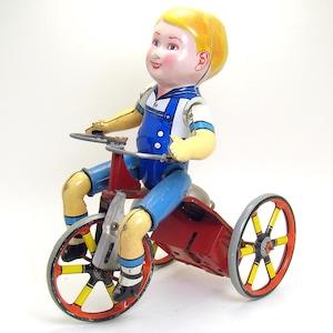 May include: A vintage tin toy of a boy riding a red tricycle with yellow wheels. The boy is wearing a blue and white sailor shirt and blue overalls. The toy has a wind-up key on the side.