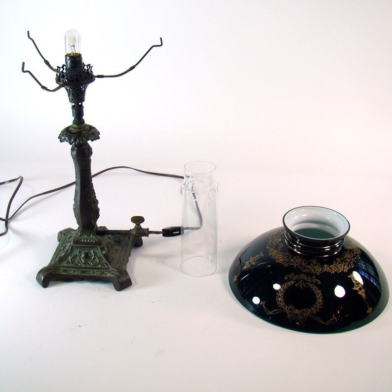 Puede incluir: Una l&aacute;mpara vintage de metal verde con una chimenea de vidrio transparente y una pantalla de vidrio negro con detalles dorados. La l&aacute;mpara tiene una base decorativa y un brazo de metal que sostiene la bombilla.