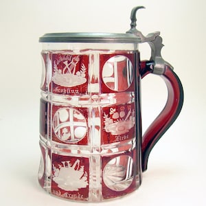 May include: A vintage glass beer stein with a red and clear paneled design. The stein features etched white designs and text, a red handle, and a metal hinged lid. The text includes the words "Frohsinn", "Liebe", and "und Freude".
