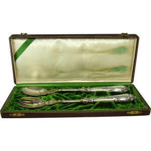 May include: A set of silver salad servers in a brown velvet-lined box. The servers have ornate handles with floral designs and are in excellent condition.