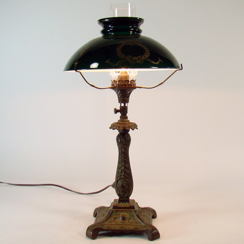 Puede incluir: Una pantalla de l&aacute;mpara de vidrio verde con detalles dorados se asienta sobre una base de l&aacute;mpara de bronce con un dise&ntilde;o ornamentado. La l&aacute;mpara tiene una chimenea de vidrio transparente y un cable.