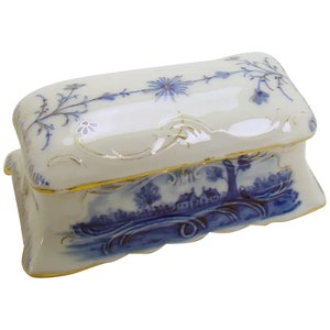 May include: A white porcelain trinket box with a blue and gold floral design. The box has a hinged lid and a decorative, scalloped edge.