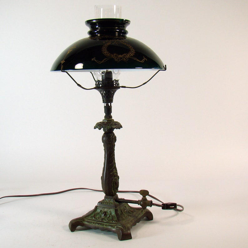 Puede incluir: Una pantalla de l&aacute;mpara vintage de vidrio verde con detalles dorados se asienta sobre una base de l&aacute;mpara de bronce con detalles ornamentados. La l&aacute;mpara tiene una chimenea de vidrio transparente y un quemador de metal.