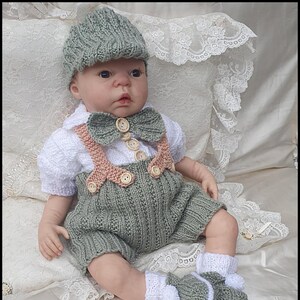 Puede incluir: Un conjunto de bebé tejido en verde salvia, con gorro, pajarita, tirantes y pantalones cortos. El conjunto incluye botines a juego con ribete blanco y pompones. Botones de madera adornan el conjunto. El texto "Pattern E61 © Babydoll Handknit Designs 2025" está en la parte inferior.