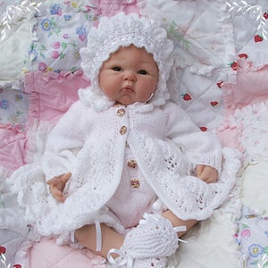 May include: A white crocheted baby doll outfit with a matching bonnet and booties. The outfit features a lacy cardigan with a button closure and a ruffled skirt. The doll is lying on a pink and white patchwork quilt.