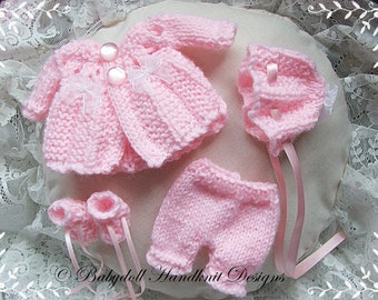 May include: A pink knitted baby doll outfit with a jacket, pants, booties, and a hat. The outfit is decorated with white bows and pearl buttons.