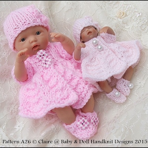 Może przedstawiać: Dwa dzianinowe stroje dla lalek niemowlęcych w kolorze różowym z białymi akcentami. Stroje obejmują sukienki, czapki i buciki. Sukienki mają wzór warkoczowy i są ozdobione guzikami w kratkę. Buciki mają kratkowe akcenty i perłowe ozdoby.