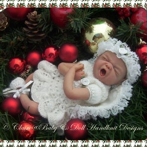 Könnte beinhalten: Ein schlafendes Baby-Puppenkleid aus weißem Häkelstoff mit einer Spitzenborte, getragen von einer schlafenden Baby-Puppe, umgeben von roten und grünen Weihnachtsdekorationen.