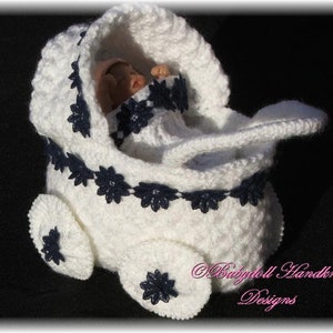 Könnte beinhalten: Weißer gehäkelter Babywagen für eine schlafende Puppe. Der Wagen hat eine blaue Spitzenverzierung und Räder. Der Text "Babydoll Handknit Designs" ist unten rechts sichtbar.