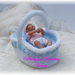 Op de afbeelding: Een witte gehaakte babymand met blauwe rand en een kleine babypop in een roze jurk.