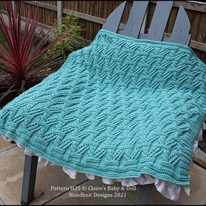 May include: A teal blue knitted baby blanket with a cable stitch pattern. The blanket is draped over a wooden chair with a white ruffle trim. The text "Pattern H25 © Claire's Baby & Doll Handknit Designs 2021" is visible at the bottom of the image.
