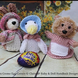 May include: Three knitted Easter egg covers, a bunny, a duck, and a bear, with a Creme Egg chocolate egg in the foreground. The bunny is wearing a pink and white dress, the duck is wearing a blue and white dress, and the bear is wearing a pink and white dress. The egg is wrapped in purple foil with gold lettering.