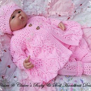 May include: A baby in a hand-knitted pink outfit, comprising a hat, jacket, and trousers. The outfit has detailed patterns and is decorated with buttons and small pearl embellishments. The baby is lying on a pink floral blanket.