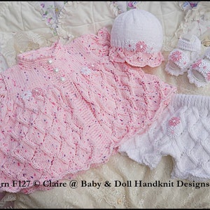 Könnte beinhalten: Ein rosa gestricktes Babykleid mit weißen Verzierungen und Perlknöpfen, eine weiße gestrickte Mütze mit rosa Verzierungen und Blumen, weiße gestrickte Shorts mit rosa Blumen und weiße gestrickte Schuhe mit rosa Blumen. Muster F127 © Claire @ Baby & Doll Handknit Designs 2017