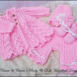 Puede incluir: Un conjunto de ropa de bebé rosa hecho a mano, que incluye una chaqueta con tres botones, un gorro, patucos y pantalones. El conjunto está tejido a ganchillo con un punto texturizado y adornado con cintas blancas. El conjunto se muestra sobre una superficie de encaje blanco.
