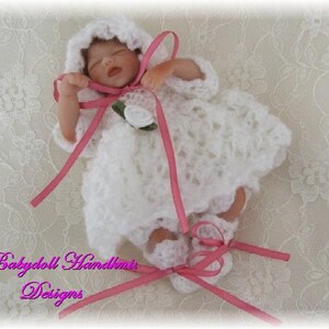 May include: A small, white crocheted dress with pink ribbon accents and a white flower. The dress is worn by a porcelain baby doll with closed eyes and a pink ribbon in her hair.