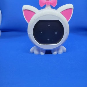 May include: White cat-shaped speaker cover with pink ears and a pink bow on top. The speaker cover is designed to fit a small, round speaker with buttons.