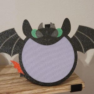 Op de afbeelding: Een zwarte en groene glitterdraak met een paarse viltlichaam en een oranje viltstaart. De draak heeft een witte viltcirkel in het midden om op te schrijven of te tekenen.