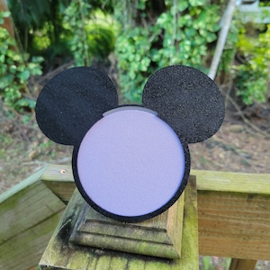 Puede incluir: Un altavoz con forma de Mickey Mouse de color morado y negro con una cubierta de tela.