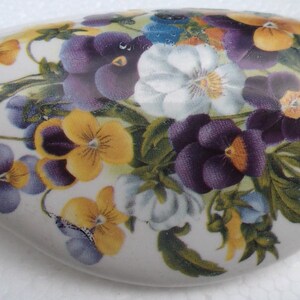 May include: A white ceramic heart-shaped trinket box with a floral design featuring purple, yellow, and white pansies. The pansies are surrounded by green leaves.