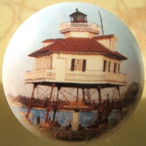 May include: A white and brown lighthouse with a red roof stands on stilts over water. The lighthouse has a white railing and windows. The image is a ceramic knob.