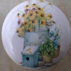 May include: A ceramic knob with a painted design of a birdhouse, flowers, and a bird. The birdhouse is blue with a white roof and a small opening. The flowers are yellow with brown centers. The bird is brown with a yellow beak. The background is white.