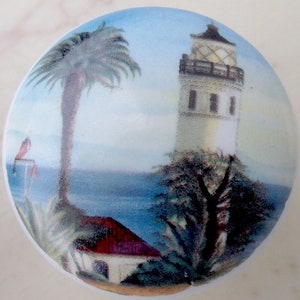 May include: A round ceramic knob with a painted image of a lighthouse, palm trees, and a blue ocean. The lighthouse is white with a black roof and windows. The palm trees are green and the ocean is blue.