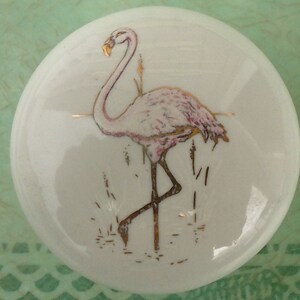 May include: White ceramic knob featuring a pink flamingo with gold accents. The flamingo is depicted standing in water with reeds. The knob has a rounded shape and is likely for a cabinet or drawer.