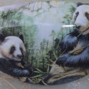 May include: White ceramic decorative drawer pull featuring a detailed illustration of two pandas in a bamboo forest. The pandas are black and white, with lush green foliage in the background. The pull is oval-shaped.