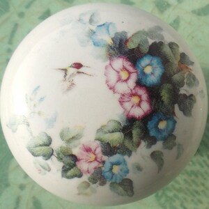 May include: A white, round ceramic knob featuring a floral design with pink, blue, and white morning glories and green leaves. A hummingbird is also depicted. The knob is likely for a cabinet or drawer.