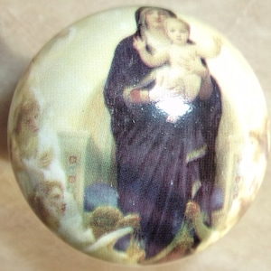 May include: A round, white, and gold-toned ornament with a painted image of a woman in a dark blue robe holding a child. The woman and child are surrounded by a light gold background.