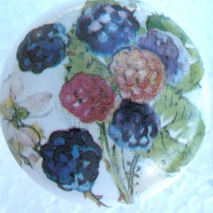 May include: A white, round decorative knob featuring a floral design. The design includes a bouquet of flowers in shades of purple, red, and orange, with green leaves. The knob is likely made of ceramic or porcelain.